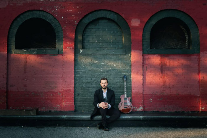 A color portrait of a musician posing in front of a red wall in Brooklyn